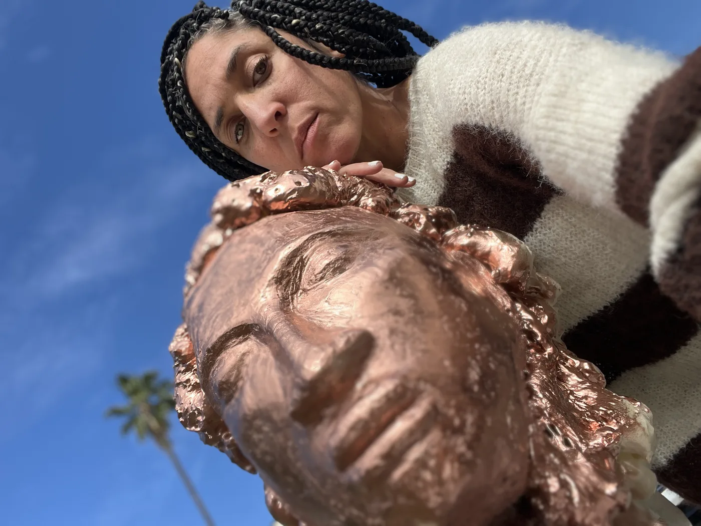 Maryam leaning over a copper and gold sculpted head against a blue sky