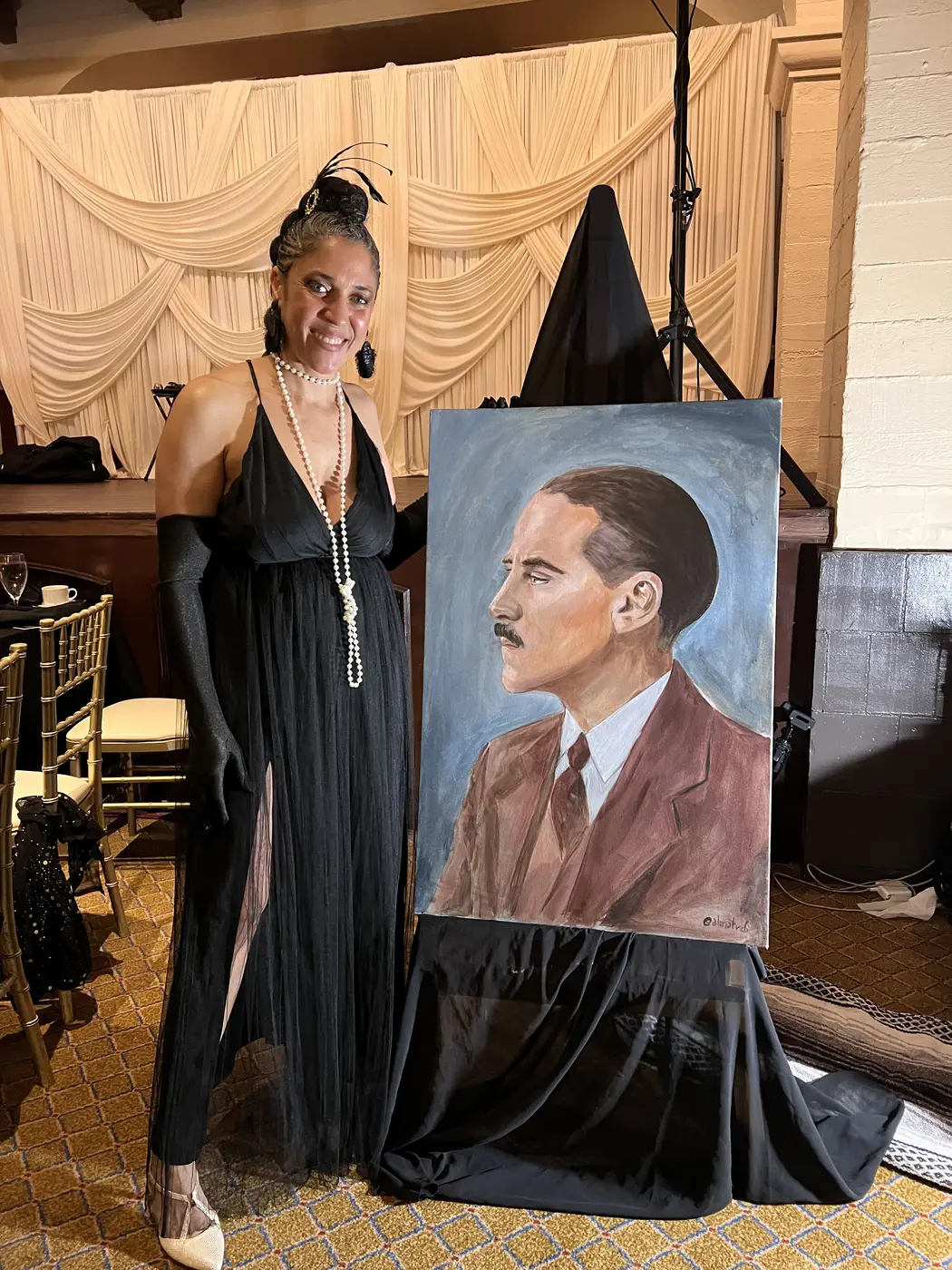 Maryam at an event beside her acrylic portrait of a man in a suit, displayed on an easel