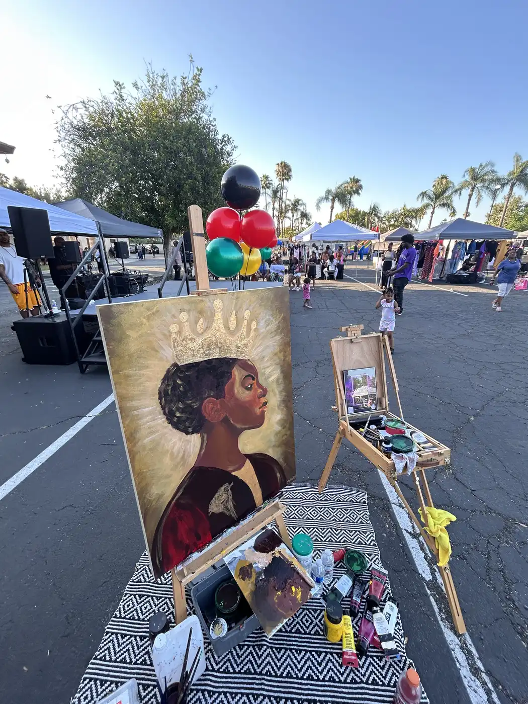 The crowned portrait on its easel beside Maryam's paints, with the event tents behind