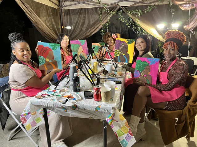 Five women in pink aprons painting portraits at a table under an evening tent