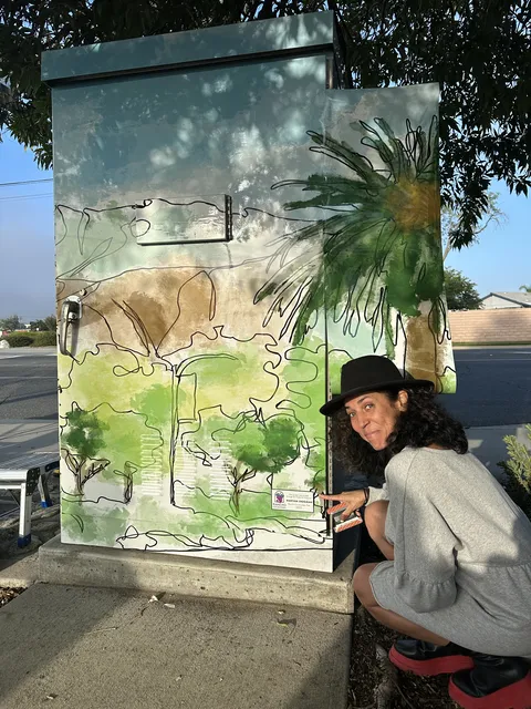 Maryam crouching beside a Rancho Cucamonga utility box she painted with trees and hills